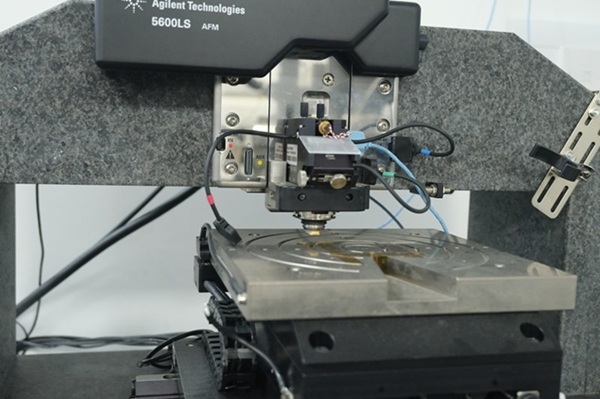 a precision scientific instrument setup, likely an Atomic Force Microscope (AFM). The main body of the microscope has a dark upper section. Below it is a metallic panel with embedded screws, wires, and technological components.