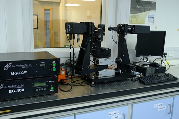 laboratory setup featuring a sophisticated piece of equipment resting on a perforated metal table. Central to the setup is a black electronic device consisting of two vertical supports connected by a series of horizontal platforms and components, likely used for scientific measurement or research.