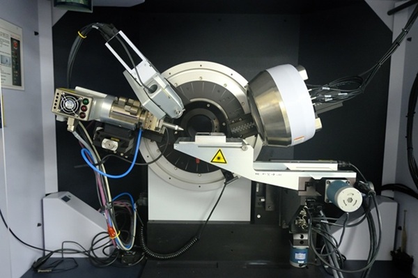 scientific instrument, likely an X-ray diffractometer, within a laboratory setting. At the center is a circular component with a black and white design, flanked by mechanical arms and electronic attachments.