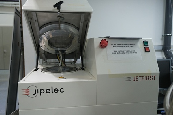 A laboratory machine with an open chamber, branded as "Jipelec" and "JETFIRST," showing its metallic interior and featuring an emergency stop button. The top section of the machine is open, revealing a circular metallic part with a glass section at the centre.