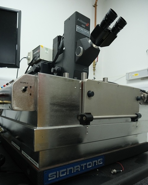 Piece of laboratory equipment, specifically a microscope setup with dual eyepieces mounted on a metallic platform and placed in a lab environment. The equipment features several knobs and controls.