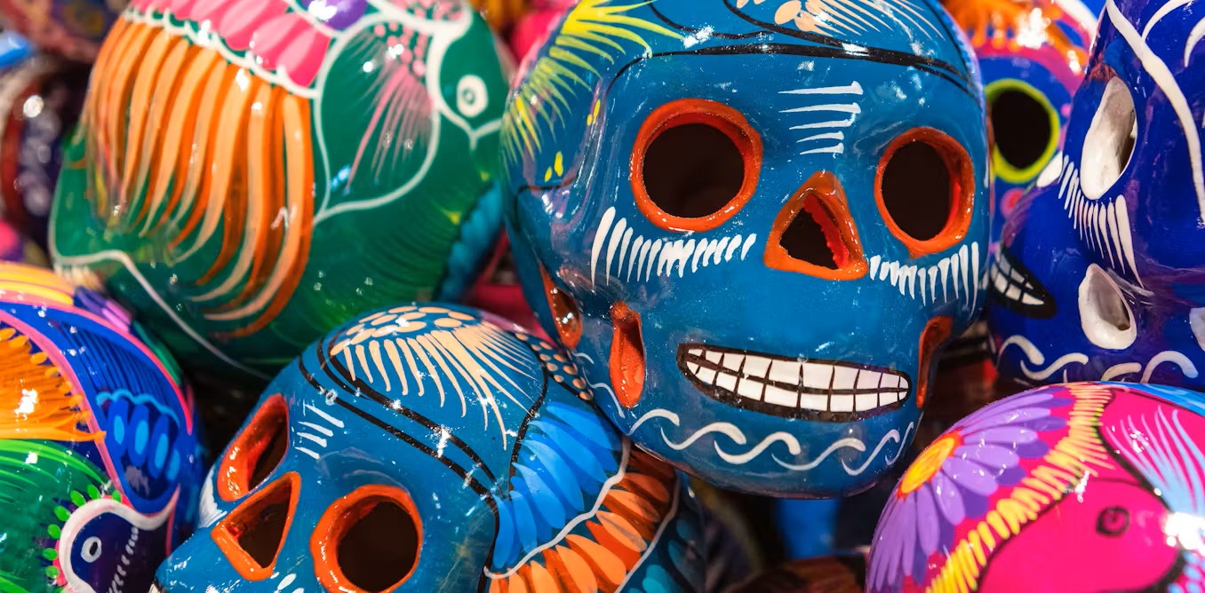 A close-up of colorful ceramic skulls decorated with vibrant patterns in blue, orange, pink, and green, commonly associated with Day of the Dead celebrations. The skulls are stacked closely together, showcasing intricate painted details.