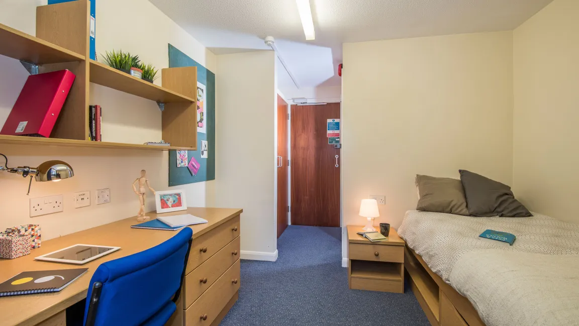 A student bedroom containing a working desk area and a neatly made bed. In the background a door opens to a bathroom.