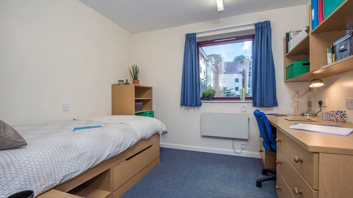 A window lets light into a bedroom where a neatly made bed and desk can be seen.