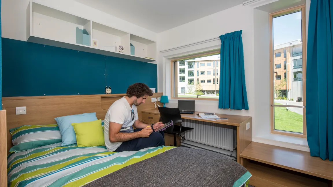 A student sits using a tablet on a large, brightly coloured bed in a modern bedroom. Modern buildings and a grassy courtyard can be seen through the windows.