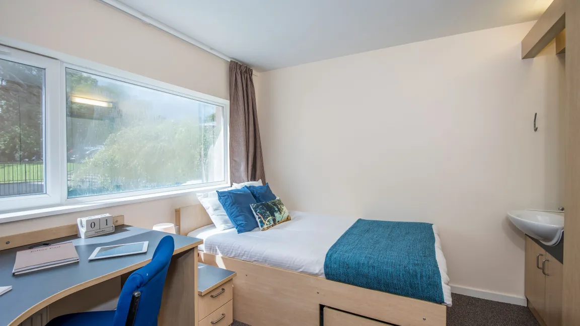 A large window lets light into a bedroom where a neatly made double bed, wash basin and desk can be seen.