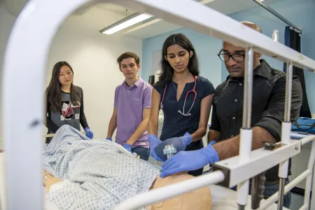 A group of students watch as one of them works on a dummy patient in a hospital bed