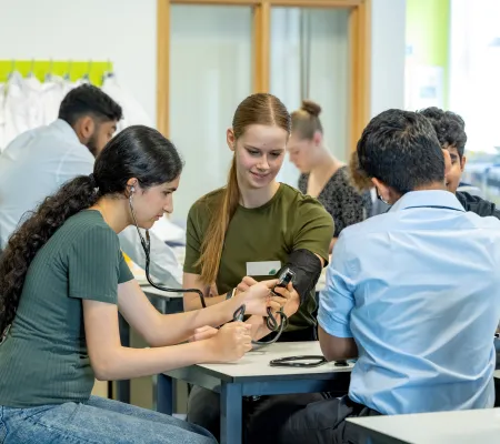  A small group of students sitting around a table watching one person measuring another’s blood pressure using a manual cuff and stethoscope, under peer observation.