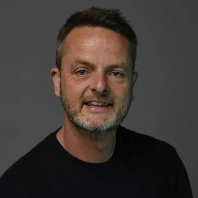 Headshot of a smiling man with short dark hair and a beard, wearing a black top against a dark grey background.