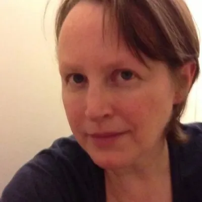 Close-up portrait of a person with short light-brown hair wearing a dark top, photographed indoors against a plain light-colored background.