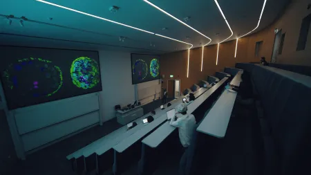 Students seated in a modern lecture theatre while a lecturer presents colourful scientific cell visualisations on large screens.