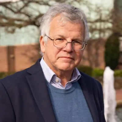 Close-up outdoor portrait of an older adult with grey hair and glasses wearing a blazer and sweater, with a blurred fountain and trees in the background.