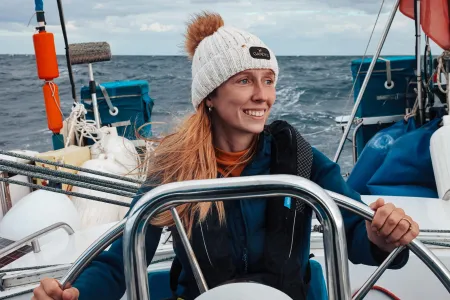 A person stands at the wheel of a small sailing boat on open water. The person wears a knitted hat with a pom-pom and a padded jacket.