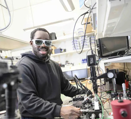 Student looking at the camera as he stands in front of a bench with lots of photonics laboratory equipment.