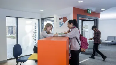 A student talks to a receptionist sat behind a desk in an apartment building lobby.