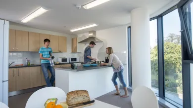 A group of students relax and talk whilst cooking food in a large, modern communal kitchen.