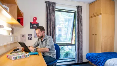 A student sits looking at a tablet computer while working at a desk. A bed and wardrobe can be seen behind him, an open window lets light into the room.