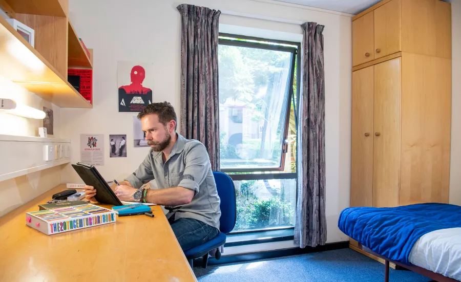 A student sits looking at a tablet computer while working at a desk. A bed and wardrobe can be seen behind him, an open window lets light into the room.