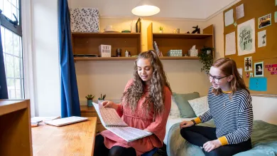 Two students sat in a bedroom talking and looking at books and notes. Light streams in through a large window.