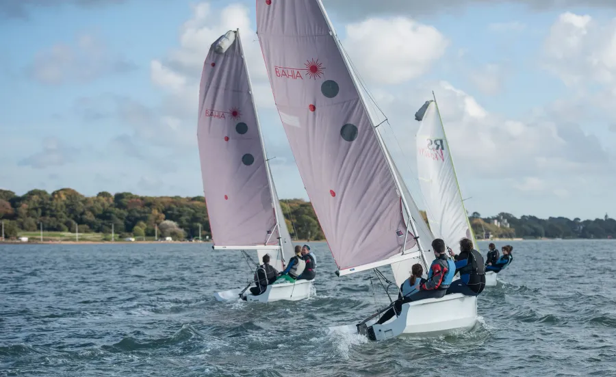 Three small sailing boats, each with a crew of three, on open water beneath blue skies. An area of trees is visible in the background.