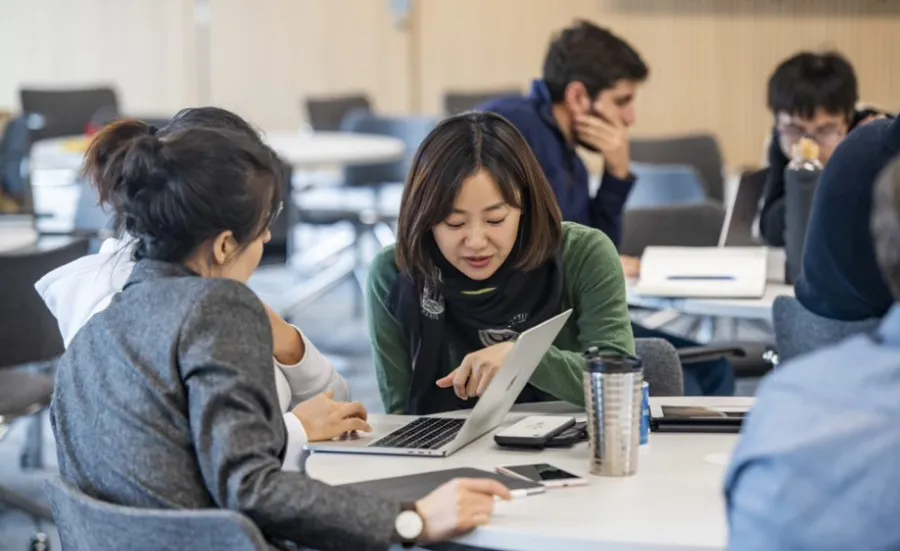 A small group of students sitting around a table look at a laptop screen and discuss their work.