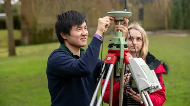 Two students conduct an experiment outdoors using a large piece of survey equipment on a tripod.