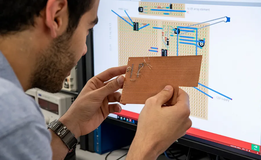 A student compares a prototype PCB to his design on a computer monitor behind it.