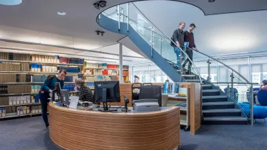 A bright, modern library reception area with shelves full of books. A lady at the desk helps someone. Two students descend a spiral staircase from an upper floor.
