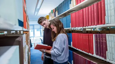 Two students in a narrow library aisle, surrounded by shelves of books on both sides. They are reading a book pulled from one of the shelves.