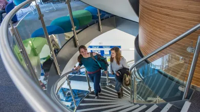 Two students ascend a set of spiral stairs from a lower to an upper floor. Study spaces, desks and stools are visible on the upper floor.