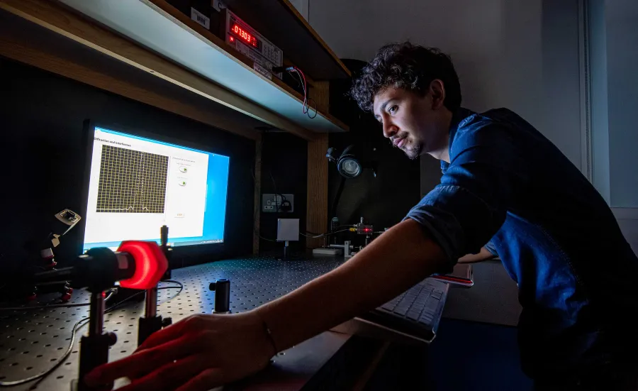 A student conducts an experiment in a dark laboratory, using a computer and some technical equipment on a large desk.