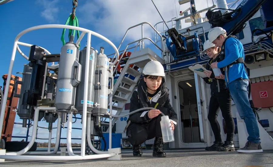 Three students outside on the deck of a boat, on a sunny day. One crouches down to collect a water sample from a large piece of technical equipment.