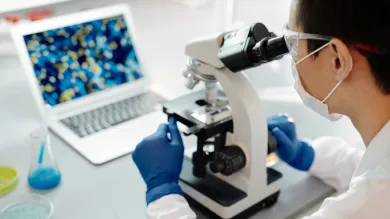 Student in a lab coat is looking into a microscope and showing results on the laptop infront of her