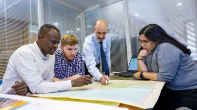 A Southampton Geospatial researcher talks with team members, standing over a map, with a laptop with a data visualisation to the side