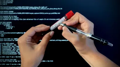 Close-up of hands labelling a patient blood sample for molecular analysis. In the background a computer screen shows programme code to process sequencing data. 