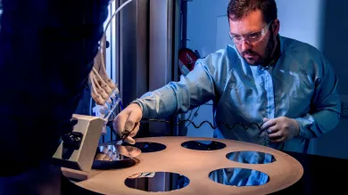 A bearded young scientist in protective overalls and glasses works with a flame hydrolysis deposition (FHD) system