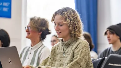 A student is sitting at a desk, typing on their laptop. They have blonde curly hair and a beige knitted jumper. Other students are sitting in the same room but are out of focus. They all face the same direction and are smiling.