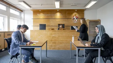 A group of 5 smartly dressed law students practise a court case in a mock courtroom.