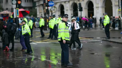 A number of police officers monitor crowds of people on a rainy London street. They are wearing high visibility yellow jackets.