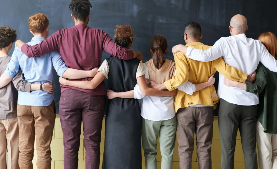 A diverse group of nine people stand in a row with their arms around each other's shoulders, facing a blackboard in a classroom, dressed in an array of casual to smart-casual attire.