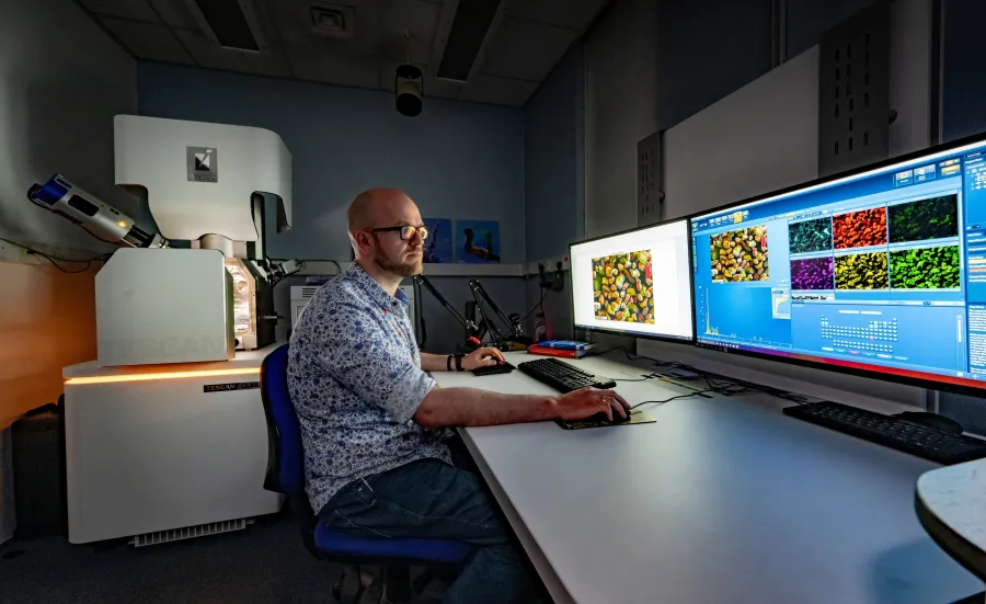 A technician works at computers displaying microscopic images, with a TESCAN electron microscope nearby.