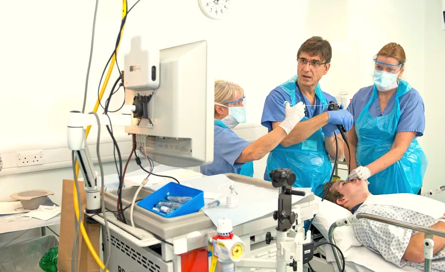 Three medical professionals conduct a procedure on a patient lying on a hospital bed, surrounded by medical equipment and supplies, in a sterile clinical environment.