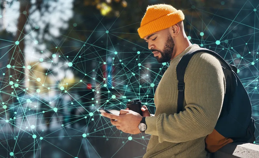 A man in a yellow beanie and olive-green sweater, engrossed in a smartphone, holding a disposable coffee cup, and wearing a backpack. He is standing against a backdrop of an abstract digital network design with interconnected nodes and lines.