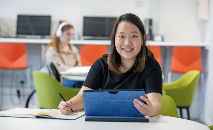Jessie is sitting at a desk smiling at the camera. She has a tablet in 1 hand and a pen and note book in the other hand.