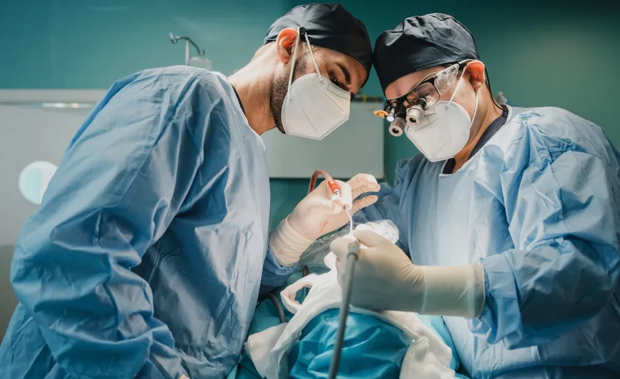 Two men in a surgery theatre. They're wearing scrubs, safety masks, and hats. They're holding a medical operating tool.  