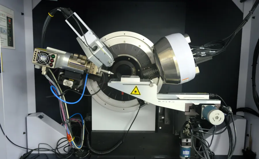 Complex scientific instrument, likely an X-ray diffractometer, set up inside a laboratory environment. It features a cylindrical component at the centre, surrounded by various metallic parts and wiring. 