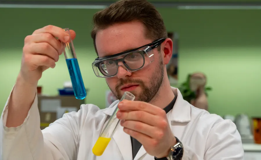 Nicholas in a lab coat and protective eyewear. He's holding two test tubes; one is filled with blue liquid, the other filled with yellow liquid.