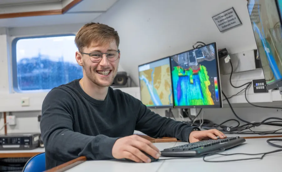 Daniel Hampton is sitting at the work station on Research Vessel Callista. There are screens displaying coulourful charts in the background.
