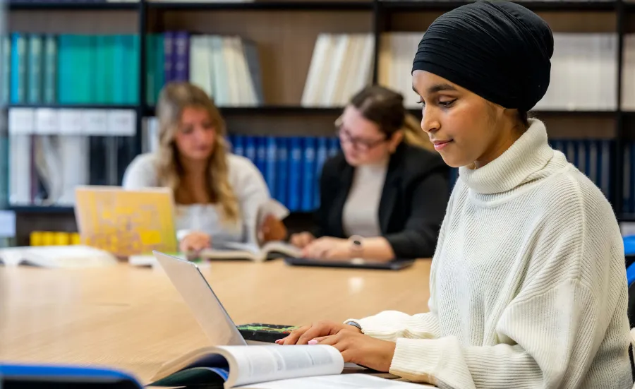 Student in a library studying with a laptop and open books at a table.