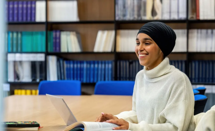 A smiling student wearing a black headscarf and cream jumper studies at a library table with books and a laptop.